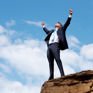 Photo of joyful businessman raising his arms upwards while standing on the rocky cliff
